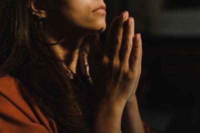 Portrait en gros plan d'une femme en prière, les mains jointes levées vers le ciel sur un fond sombre.
